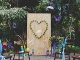 a colorful backyard wedding ceremony space with a plywood piece with a heart of greenery, bright ribbons decorating the chairs and colorful paper butterflies and blooms