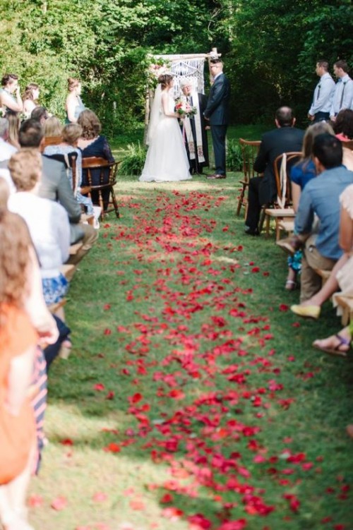 a boho backyard wedding ceremony space with red petals on the ground, a boho wedding arch with macrame is cute and cool