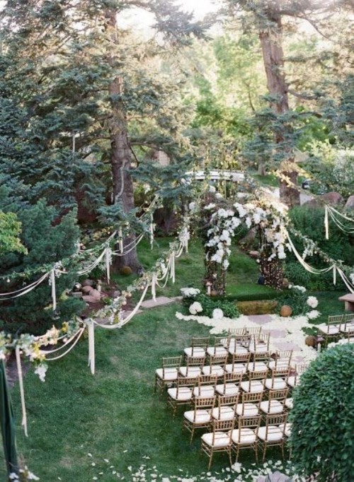 a forest backyard wedding ceremony space with a large wedding arch covered with greenery, lights and white blooms, with matching garlands all over the space adn white chairs in rows