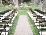 a stylish backyard wedding arch with a gravel path, white chairs decorated with pink and dark blooms and with trees for a backdrop