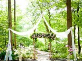 a backyard ceremony space with an arch decorated with greenery and pink blooms, with fabric straps and pink floral arrangements lining up the aisle