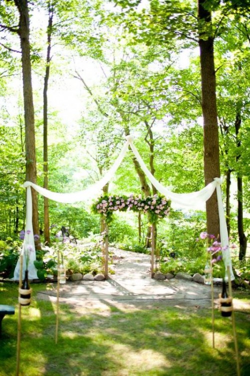 a backyard ceremony space with an arch decorated with greenery and pink blooms, with fabric straps and pink floral arrangements lining up the aisle