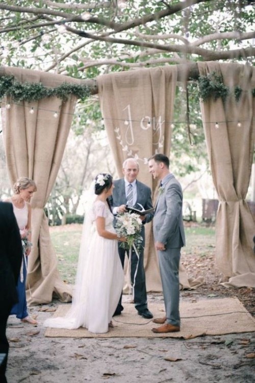 a cozy backyard rustic wedding space with burlap, greenery, lights over the space and burlap rugs
