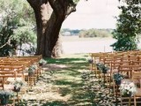 a beautiful backyard wedding ceremony space with a living tree and a lake for a backdrop, white bloom arrangements lining up the aisle and petals on the ground