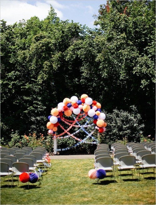 a bright and cool wedding ceremony space with white chairs in rows, an aisle marked with colorful paper balls and a wedding arch decorated with them, too