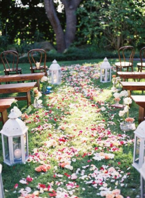 a backyard with chairs and benches, candle lanterns and bright blooms and petals right on the ground, living trees for a backdrop