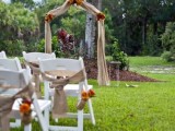 a rustic backyard wedding arch covered with burlap and sunflowers, with white chairs decorated in the same way