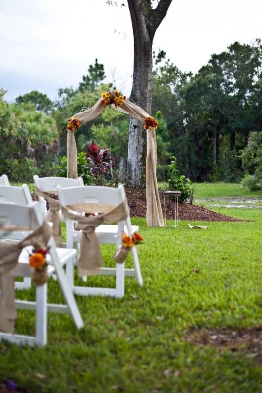 A rustic backyard wedding arch covered with burlap and sunflowers, with white chairs decorated in the same way