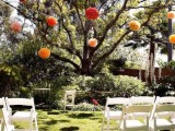 a backyard wedding ceremony space with white chairs in rows and a living tree decorated with bright paper pompoms is a lovely and cute space