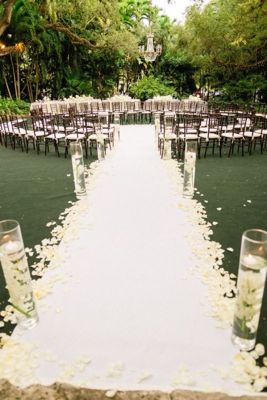 dark stained chairs with white seats and a white pathway with floating candles and petals on the path