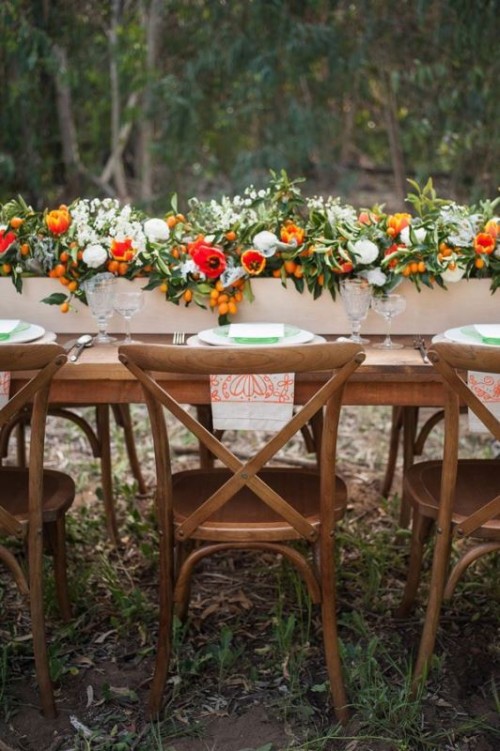 a colorful wedding tablescape with a wooden box with bright and white blooms, greenery and kumquats, white and green plates and colorful printed napkins