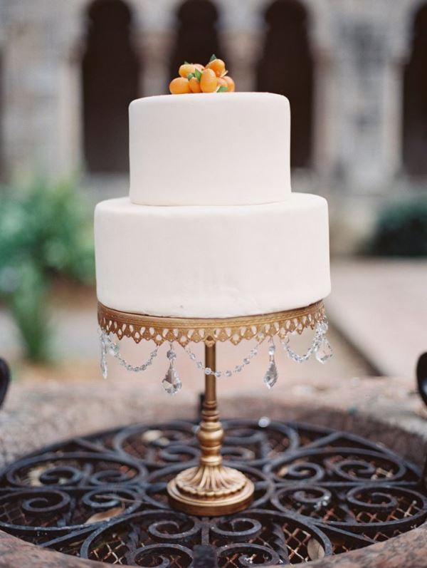 an elegant sleek white wedding cake with a bit of kumquat on top and a gold cake stand with crystals is a chic and cool idea