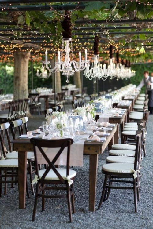 an outdoor wedding reception with a roof covered with lights and vines and with pretty crystal chandeliers that add chic to the space