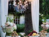 an arch with white curtains and a crystal chandelier can be used to accent a sweets table or another station of your wedding venue