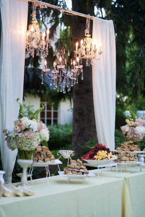 an arch with white curtains and a crystal chandelier can be used to accent a sweets table or another station of your wedding venue