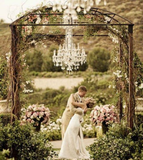 a metal wedding arbor covered with vines and blooms plus a crystal chandelier and greenery and floral arrangement is great for a garden ceremony
