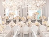 a super refined all-white and silver wedding reception with very lush florals, mirrors in ornated frames and an oversized crystal chandelier over the space