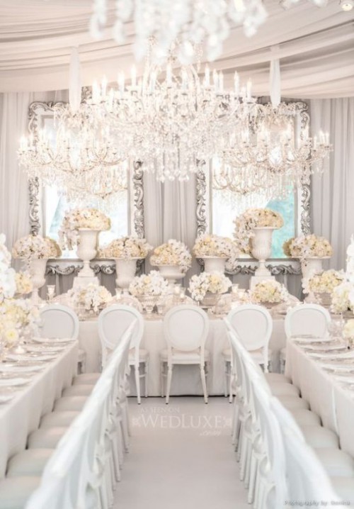 a super refined all-white and silver wedding reception with very lush florals, mirrors in ornated frames and an oversized crystal chandelier over the space