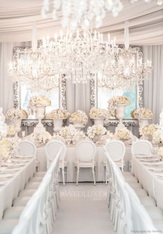 A super refined all white and silver wedding reception with very lush florals, mirrors in ornated frames and an oversized crystal chandelier over the space