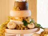 a bold and cool cheese wheel wedding cake with grapes, pumpkins and pretty doll-like toppers that look nice and cool
