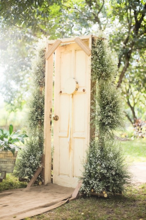 a neutral vintage wedding backdrop of a door decorated with greenery and baby's breath is a lovely idea for a rustic wedding