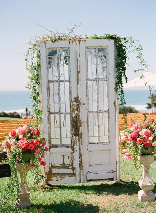 a vintage wedding backdrop of double doors topped with greenery and with neutral urns with pink blooms and greenery