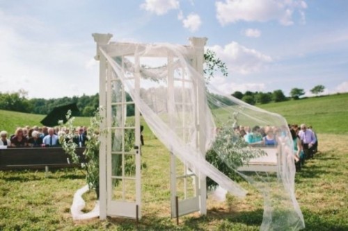 a unique wedding backdrop of a door with tulle and greenery is a lovely idea for a chic wedding outdoors