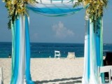 a blue beach ceremony space with a bold arch done with white and bold blue fabric, with peachy and yellow blooms and petals on the sand