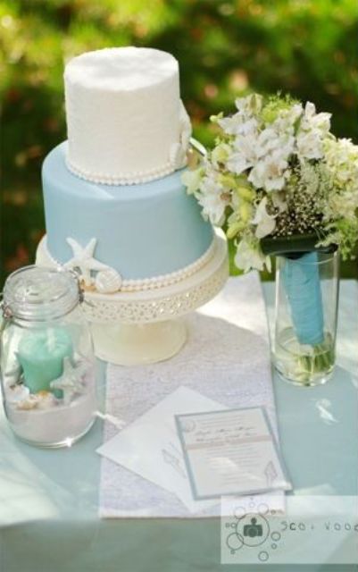 a light blue and white wedding cake with seashells and starfish plus a blue candle in a jar for a blue beach wedding