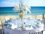 a blue and white beach wedding tablescape wiht a white orchid centerpiece, white linens, blue boxes and a gorgeous sea view