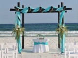 a black beach wedding arch with bright turquoise fabric, greenery and white chairs on the beach