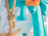 a white folding chair decorated with a turquoise ribbon, a bold bloom and a starfish hanging on rope