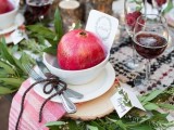 a cozy rustic wedding tablescape with a checked tablecloth, wood slices, greenery , a pomegranate in a bowl and pink checked napkins