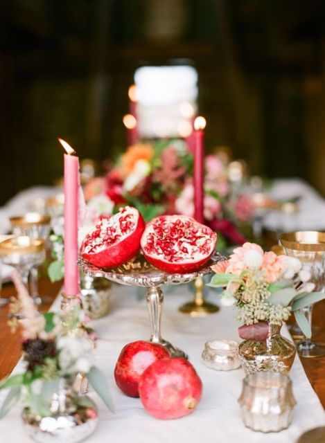 a cluster wedding centerpiece of neutral blooms, greenery, burgundy candles and a cut pomegranate on a silver stand is a gorgeous idea