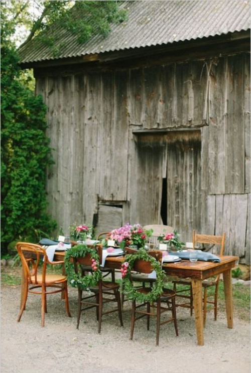 a chic rustic backyard wedding tablescape with an uncovered table, blue and white linens, pink floral centerpieces with greenery and mismatching vintage chairs with wreaths for decor