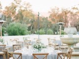 a beautiful and chic backyard wedding tablescape with a blue tablecloth, a chic white floral and greenery centerpiece, glasses and elegant stained chairs