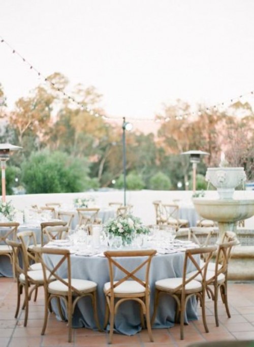 a beautiful and chic backyard wedding tablescape with a blue tablecloth, a chic white floral and greenery centerpiece, glasses and elegant stained chairs