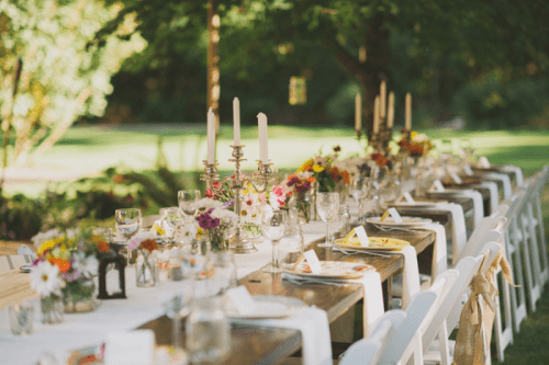 a refined backyard wedding table setting with neutral linens, candles in candle holders, bright blooms and greenery and candle lanterns is a cool idea