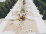 a chic backyard wedding tablescape with white linens and a burlap runner, wildflowers in vases and elegant glasses is simple and very chic