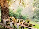 a boho backyard wedding tablescape with a rough table and benches, with neutral linens, catchy neutral and greenery centerpieces plus hanging bulbs over the space