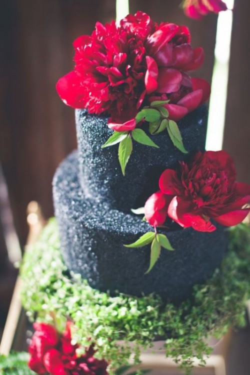 a shiny black wedding cake with bold deep red blooms and greenery is a dramatic and bold idea for a Halloween wedding, get inspired