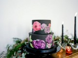 a plain black wedding cake with colorful painted blooms on it is a very chic and bold idea for a modern wedding with bright blooms