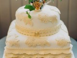a white wedding cake with neutral lace decorations and ruffles topped with a fresh bloom arrangement in a vase
