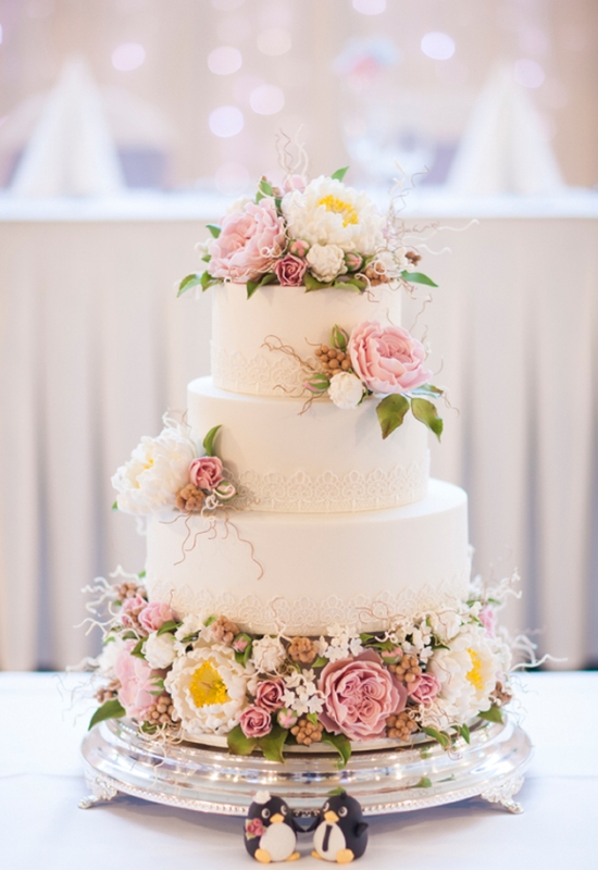 A white wedding cake with lace decor and lots of fresh blooms in pink and white and greenery