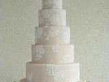 a tan wedding cake with white lace decor and pink blooms and greenery on top for a chic look