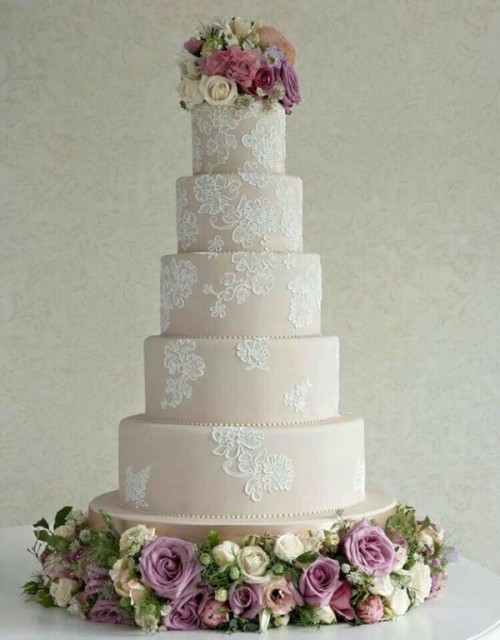 a tan wedding cake with white lace decor and pink blooms and greenery on top for a chic look