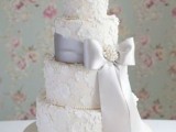 a white wedding cake with white floral lace appliques made of sugar, a lilac ribbon with a bow and a brooch and white blooms on top