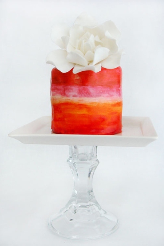 a super bright orange, yellow and pink painted wedding cake with a white sugar bloom on top is amazing for a colorful wedding