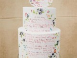 a white buttercream wedding cake with hand painted flowers and calligraphy with quotes that mean a lot to the couple plus a matching cake topper