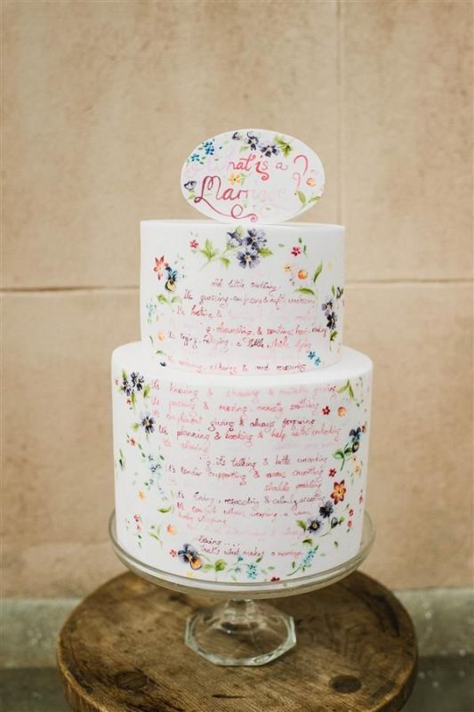 a white buttercream wedding cake with hand painted flowers and calligraphy with quotes that mean a lot to the couple plus a matching cake topper